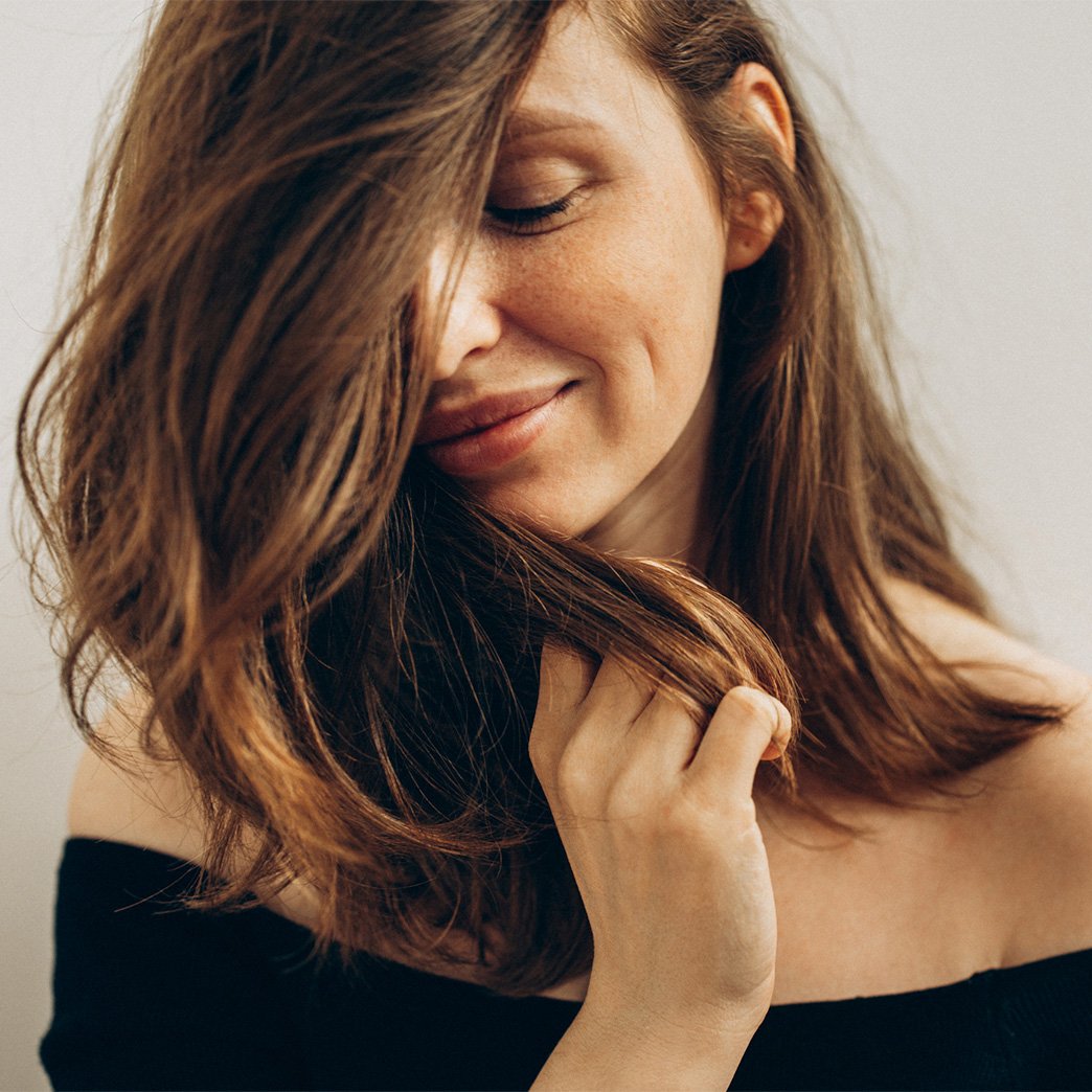 Beautiful woman with eyes closed as she hides half her face behind her smooth, wavy hair. She smiles embracing the softness.